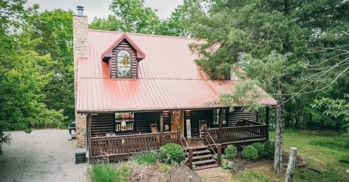 A charming log cabin with a red metal roof, surrounded by greenery and featuring a welcoming porch.