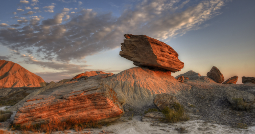A large, balanced rock sits atop a sandy landscape, illuminated by warm sunset light against a cloudy sky.