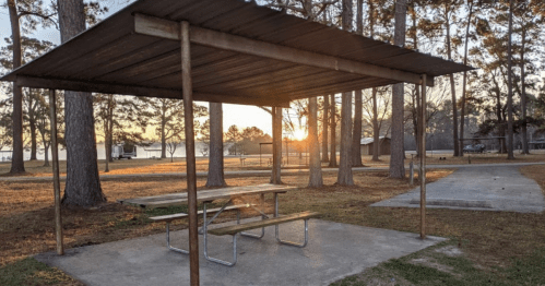A picnic shelter with a table at sunset, surrounded by trees and a calm lake in the background.