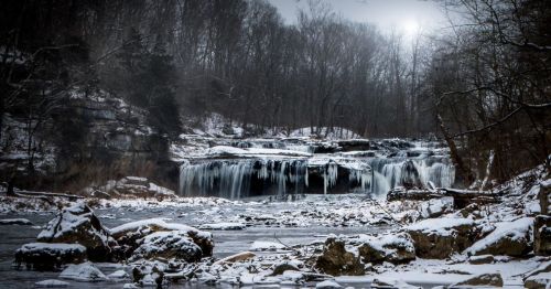 A serene winter scene featuring a frozen waterfall surrounded by snow-covered rocks and bare trees.