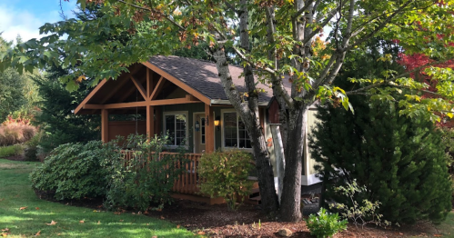 A cozy cabin surrounded by trees and greenery, featuring a porch and autumn foliage.