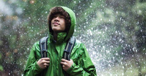 A person in a green rain jacket stands outdoors, looking up with a serene expression as rain falls around them.