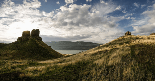 Scenic landscape featuring grassy hills, ruins, and a lake under a cloudy sky.