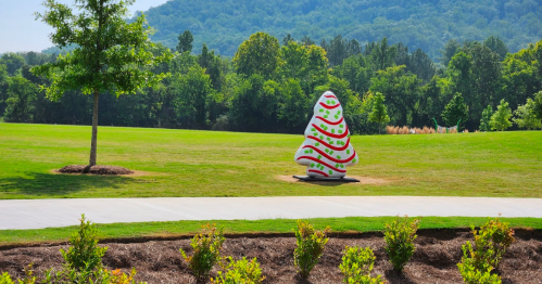 A colorful Christmas tree sculpture stands on a grassy field with trees and mountains in the background.