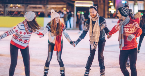 Four friends ice skating together, wearing festive sweaters, smiling and holding hands on a rink.