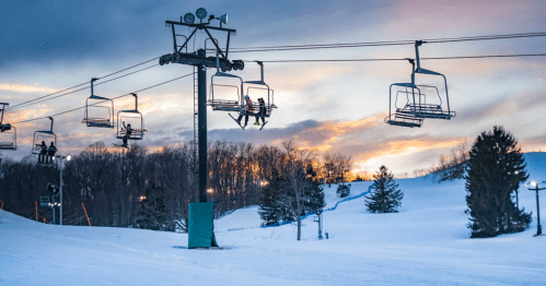 Ski lift chairs ascend a snowy slope at sunset, with trees and a colorful sky in the background.