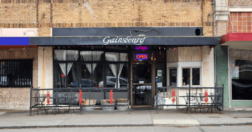 Exterior of Gainsbourg restaurant with an "Open" sign, featuring a patio and decorative elements on a brick building.