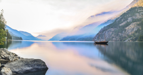 A serene lake surrounded by misty mountains, with a sailboat anchored peacefully in the calm water.