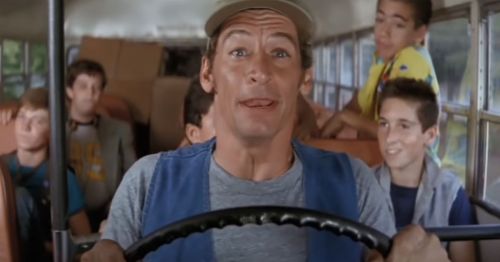 A bus driver smiles while driving, with children seated behind him, enjoying a fun ride.