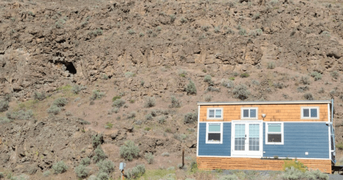 A small blue and wood-paneled house sits beside a rocky hillside covered in sparse vegetation.