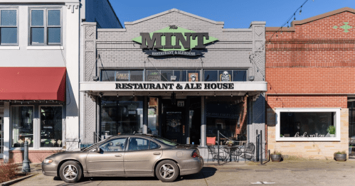 Exterior of The Mint Restaurant & Ale House, featuring a gray building with a sign, outdoor seating, and a parked car.