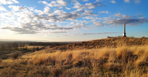 A grassy hill under a blue sky with clouds, featuring a tall monument in the distance.