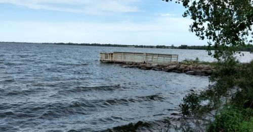 A wooden dock extends into a calm lake, surrounded by trees and a cloudy sky. Waves gently ripple on the water's surface.