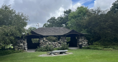 A stone pavilion surrounded by greenery under a partly cloudy sky, with a circular stone bench in front.