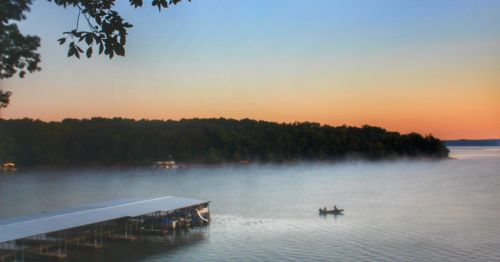 A serene lake at dawn with mist rising, a small boat gliding through the water, and trees lining the shore.