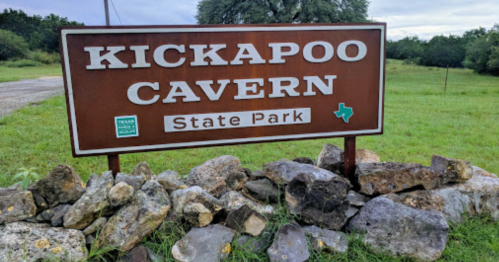 Sign for Kickapoo Cavern State Park, featuring a brown background with white text, surrounded by rocks and greenery.