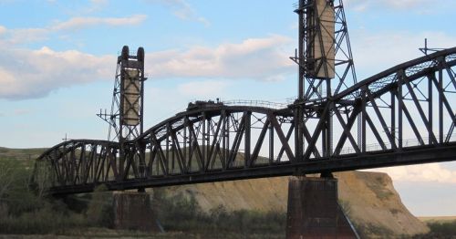 A large, metal bridge spans a river, featuring tall towers and a rugged landscape in the background.