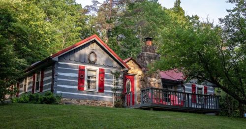 A charming log cabin with red shutters and a stone chimney, surrounded by lush greenery and a wooden deck.