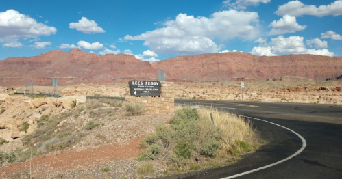 Sign for Lee's Ferry along a winding road, with red rock formations and blue sky in the background.