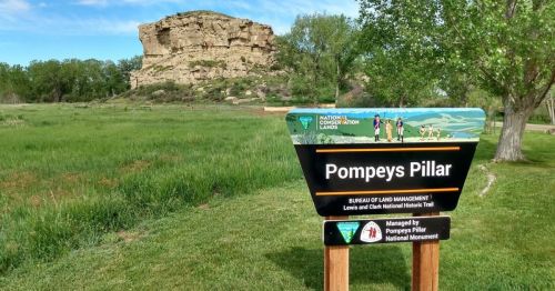 Sign for Pompeys Pillar National Monument with a rocky outcrop in the background, surrounded by green grass and trees.