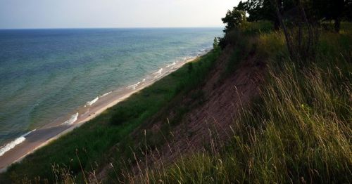 A serene beach scene with gentle waves, grassy dunes, and a clear blue sky.