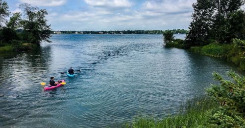 Two people kayaking on a calm lake surrounded by greenery and trees under a partly cloudy sky.
