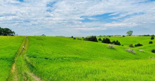 A lush green landscape with rolling hills and a dirt path, under a partly cloudy sky. Two people walk in the distance.