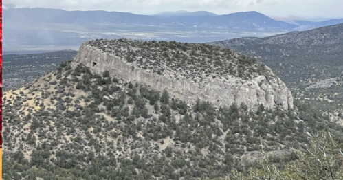 A rocky mountain peak surrounded by greenery, with distant mountains and a valley visible in the background.