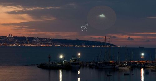 A twilight view of a marina with boats, featuring a bright object in the sky above a distant illuminated coastline.