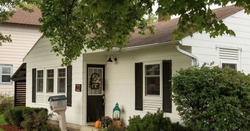 A charming white house with green shutters, a front porch, and a mailbox surrounded by shrubs and trees.