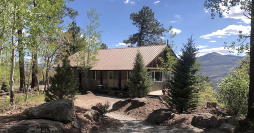 A rustic cabin surrounded by trees and rocks, set against a backdrop of mountains and a blue sky.
