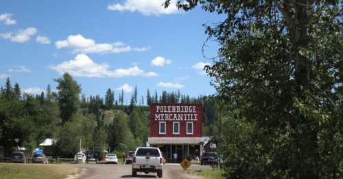 A dirt road leads to the red Polebridge Mercantile building, surrounded by trees and blue skies.