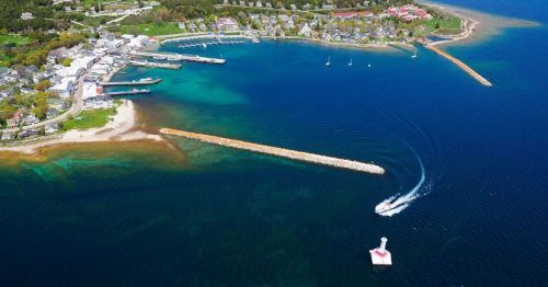 Aerial view of a coastal town with a marina, boats, and a lighthouse near a sandy beach and green hills.