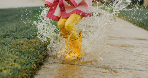 A child in yellow rain boots splashes in a puddle on a sidewalk, wearing a pink raincoat.