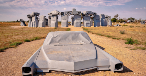 A gray car sculpture in the foreground with a stone-like structure of stacked cars in the background under a blue sky.