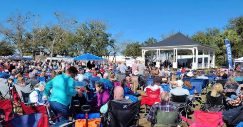 A large crowd gathers in a park for an outdoor event, with tents and a stage in the background under a clear blue sky.