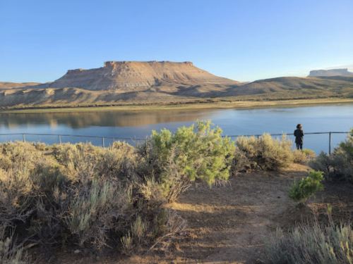 A person stands by a fence overlooking a calm river with a mountain in the background under a clear blue sky.
