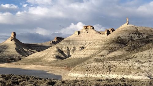 A scenic landscape featuring rugged hills and rock formations under a cloudy sky, with a calm body of water in the foreground.