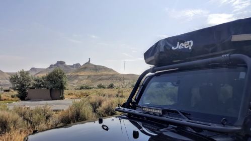 A black Jeep with a rooftop tent parked in a scenic landscape featuring hills and a distant rock formation.