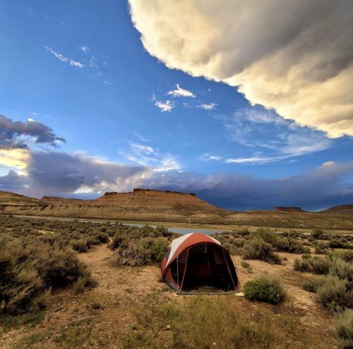A tent set up in a desert landscape with dramatic clouds and a distant plateau under a blue sky.