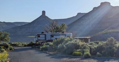 A camper parked near a scenic landscape with mountains and greenery under a clear sky.