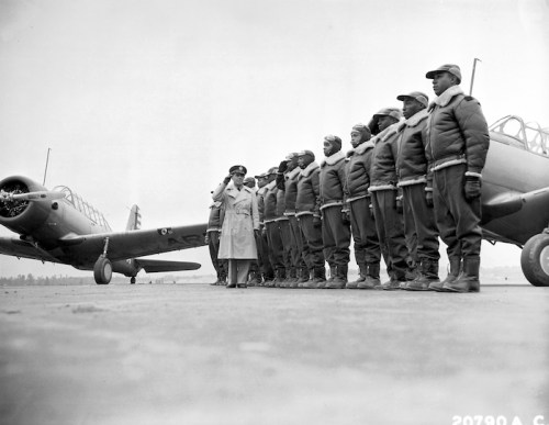 A group of uniformed soldiers stands in formation beside a vintage aircraft, with an officer inspecting them.