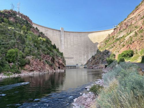 A large concrete dam rises above a calm river, surrounded by rocky hills and greenery under a clear blue sky.