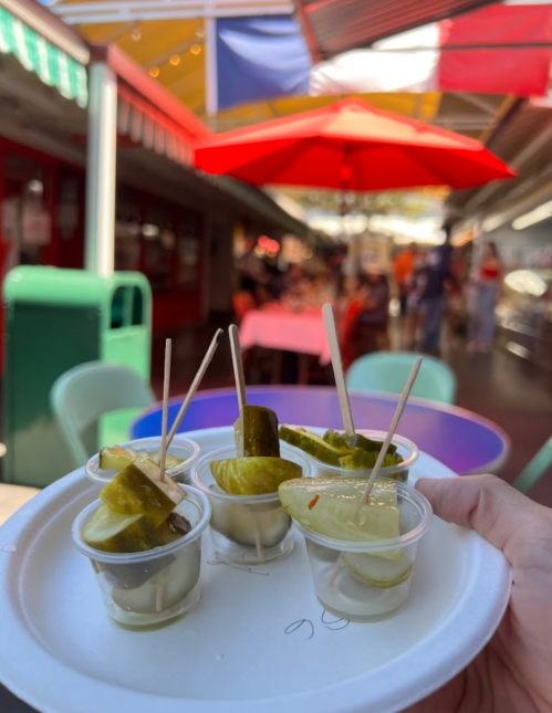 A hand holds a plate with small cups of pickles on toothpicks, set against a colorful market backdrop.