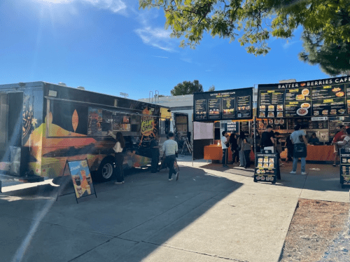 A vibrant food truck and a café stand with menus, people ordering food under a clear blue sky.