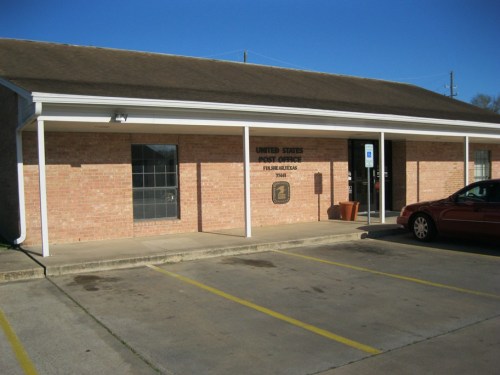 Exterior of a brick building labeled "United States Post Office" with a parking lot and clear blue sky.
