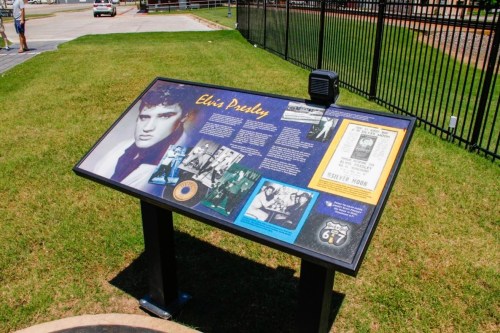 A display board featuring information and images about Elvis Presley, set on a grassy area near a fence.