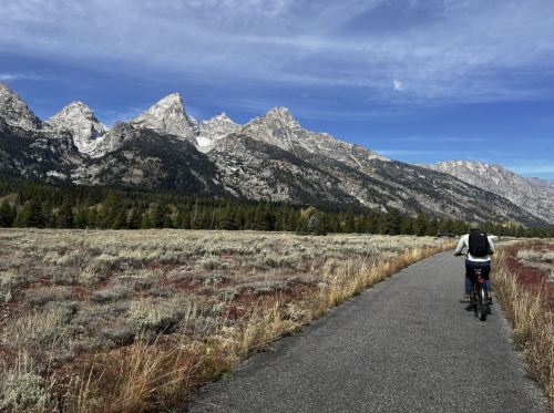 A person rides a bike on a path with mountains and a clear sky in the background, surrounded by grassy fields.