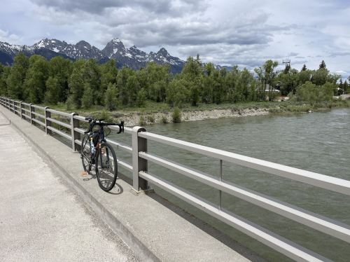 A bicycle rests on a bridge overlooking a river, with mountains and trees in the background under a cloudy sky.