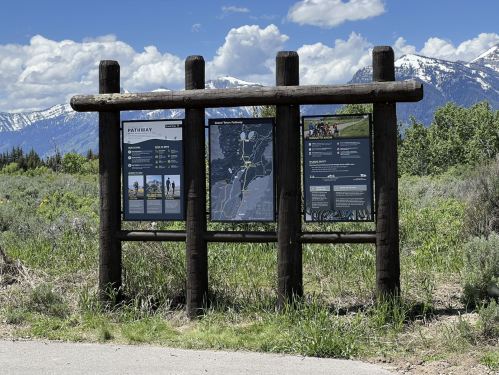 Information signpost with maps and details about pathways, set against a backdrop of mountains and blue sky.